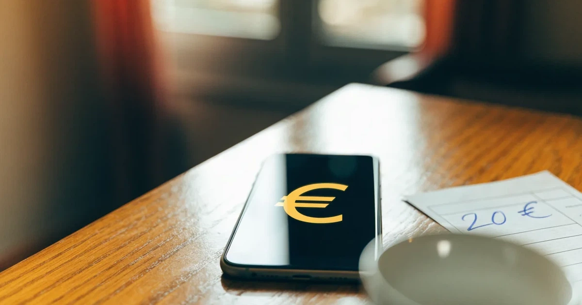 Zwei Euro-Scheine und ein Smartphone liegen auf einem Holztisch neben einer Tasse, im Hintergrund ein unscharfes Fenster.