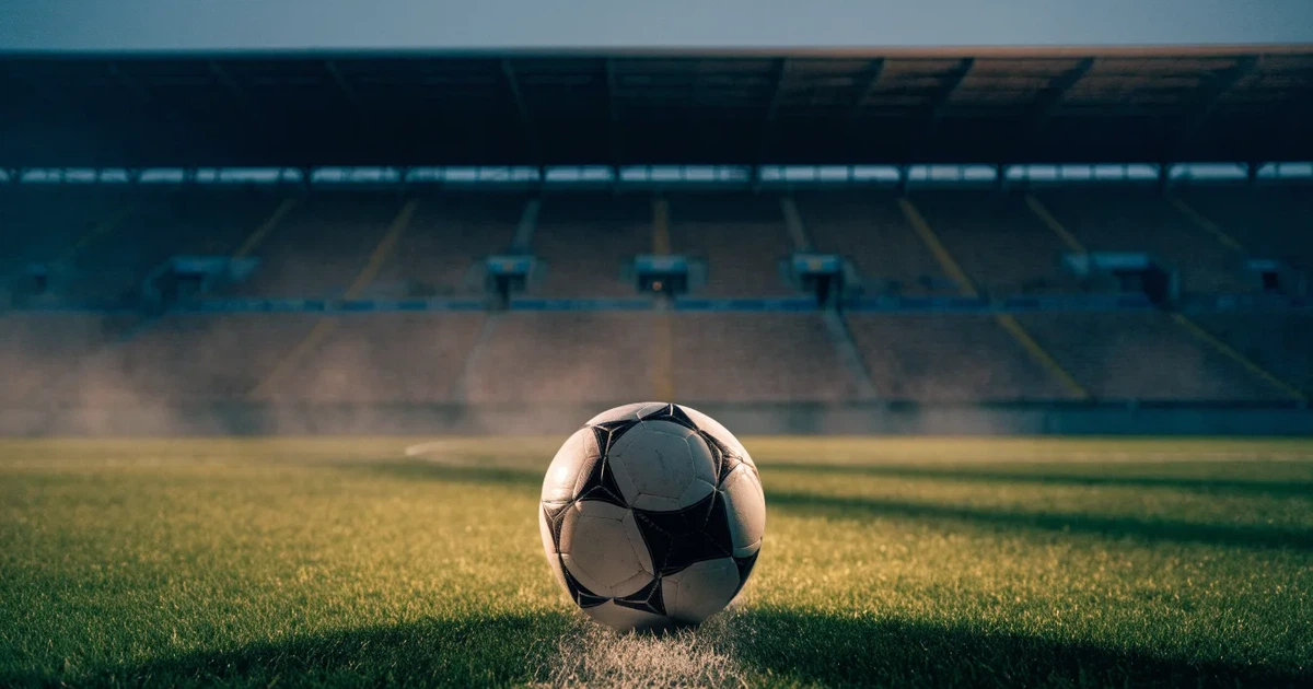Ein Fußball liegt auf dem Anstoßpunkt eines leeren Rasenplatzes, im Hintergrund eine große Stadiontribüne im Abendlicht.