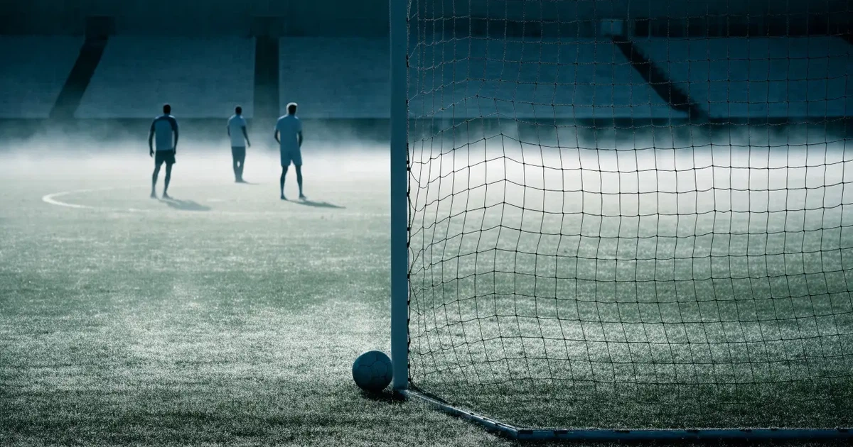 Ein Fußballplatz mit gepflegtem Rasen und einem Tor, über dem Spielfeld liegt Morgennebel, im Hintergrund eine leere Tribüne.