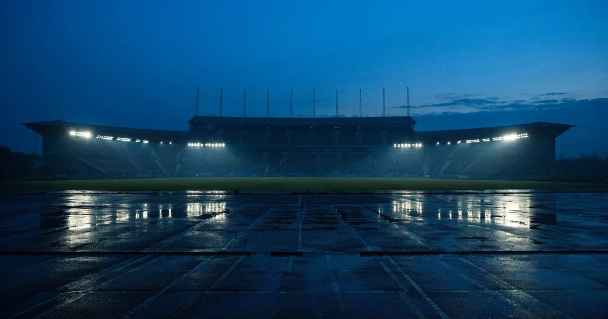 Ein Stadion unter Flutlicht aus großer Entfernung gesehen, davor ein regennasser Parkplatz in der Dämmerung.