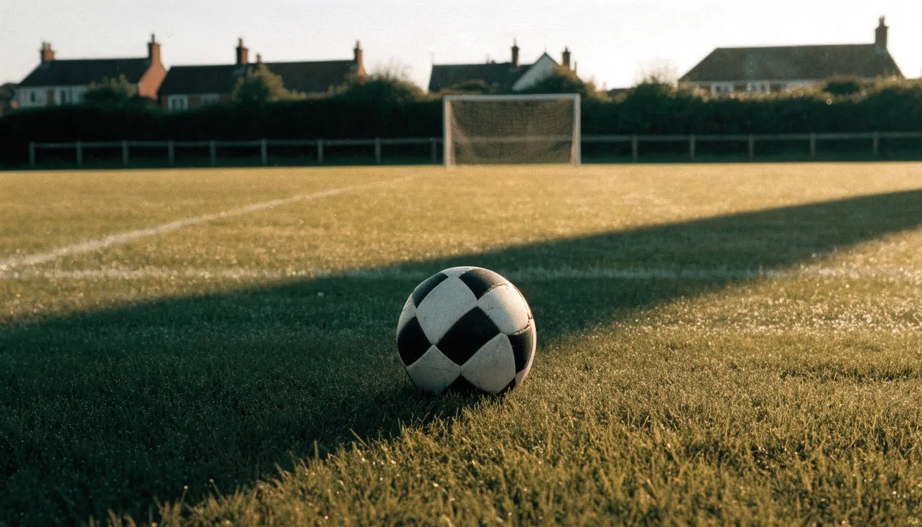 Ein Fußball auf dem Rasen eines leeren Sportplatzes am Rand einer Kleinstadt im Morgenlicht.