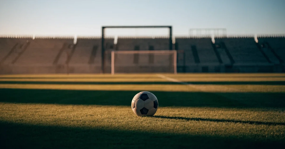 Ein Fußballplatz mit frisch gemähtem Rasen im Abendlicht, ein Ball liegt im Strafraum, im Hintergrund unscharf eine leere Tribüne.