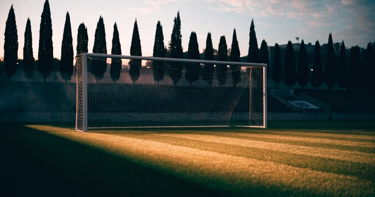 Ein Fußballtor mit Netz auf einem leeren Platz im Abendlicht, der Rasen frisch gemäht, im Hintergrund Zypressen.