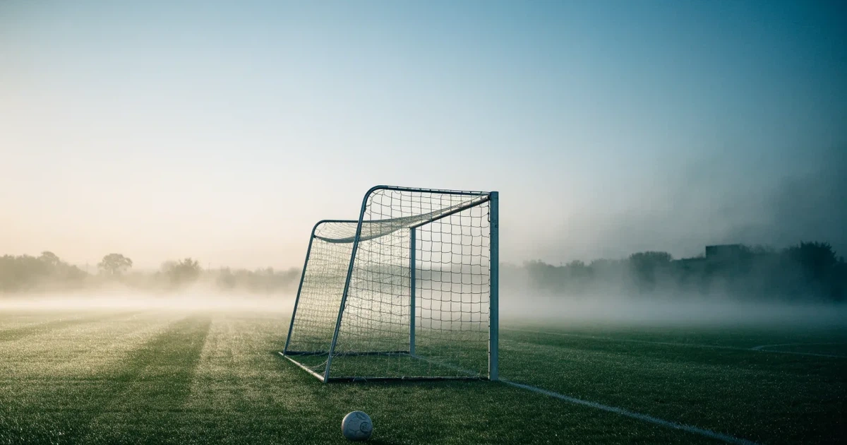 Ein Fußballtor mit Netz steht auf einem leeren Platz im Nebel der Morgendämmerung, der Rasen ist taufeucht.