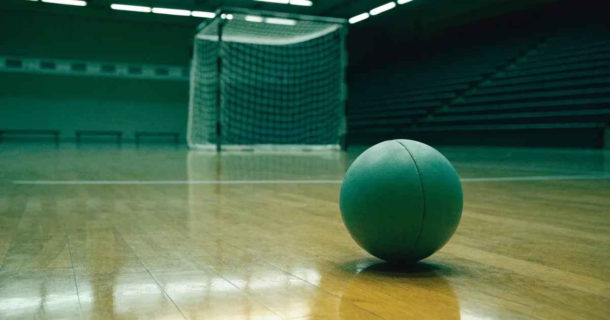 Ein Handball liegt auf dem Hallenboden vor einem Tor mit Netz, die leere Halle wird von Flutlicht beleuchtet.