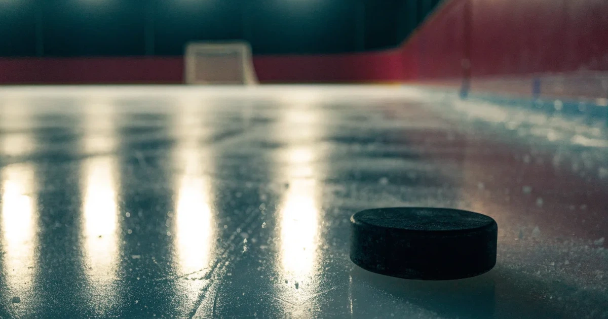 Eine leere Eisfläche in einer Halle mit Banden und Flutlicht, auf dem Eis liegt ein einzelner Puck.