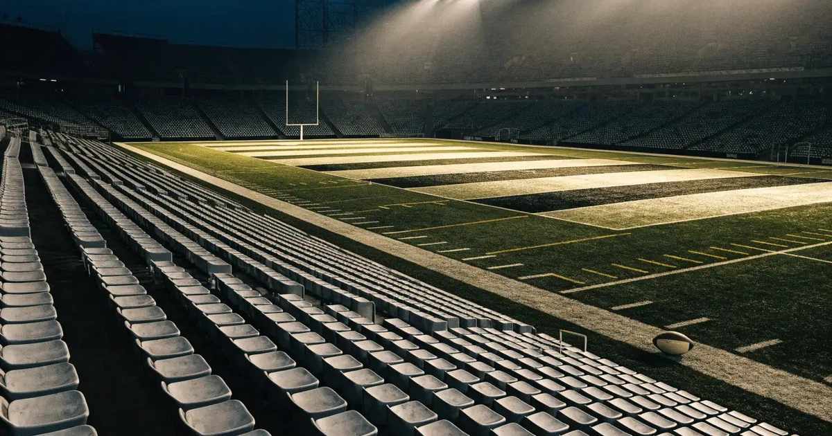 Ein Footballstadion bei Flutlicht aus der Ferne gesehen, davor ein leerer Rasen mit Yard-Linien und ein ovaler Ball am Spielfeldrand.