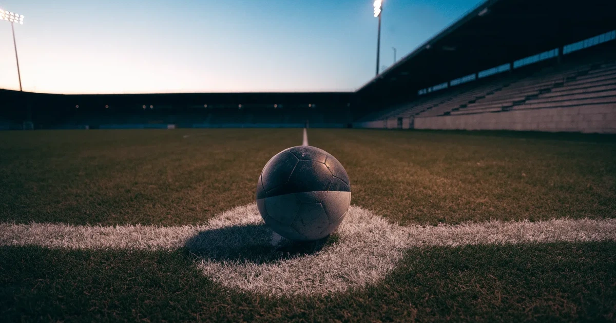 Ein Fußball liegt auf dem Anstoßpunkt eines leeren Rasenplatzes, im Hintergrund eine Tribüne und Flutlichtmasten in der Abenddämmerung.