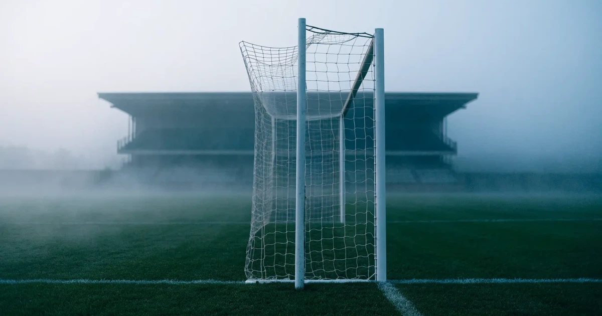 Ein Fußballtor mit Netz steht im Morgennebel auf einem leeren Rasenplatz, im Hintergrund die Silhouette einer kleinen Tribüne.