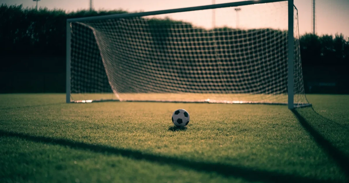 Ein Fußballtor mit Netz im Gegenlicht auf einem leeren Platz am Abend, davor ein Ball auf dem Rasen.