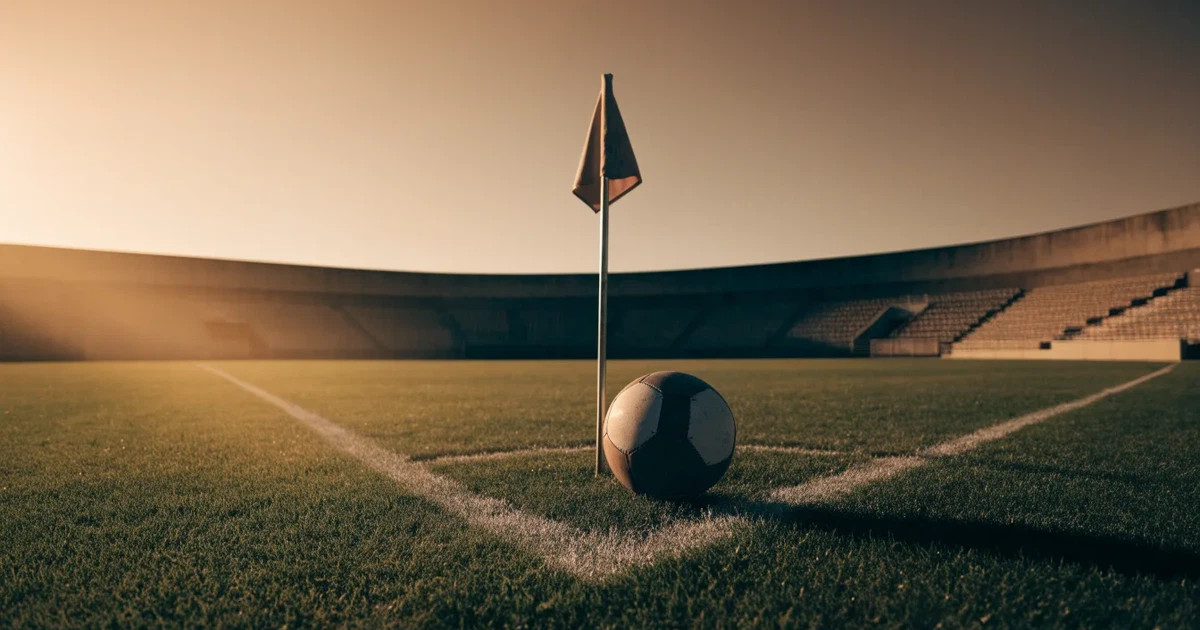 Ein Fußball liegt am Viertelkreis an der Eckfahne eines Rasenplatzes, im Hintergrund leere Tribünen in warmem Abendlicht.