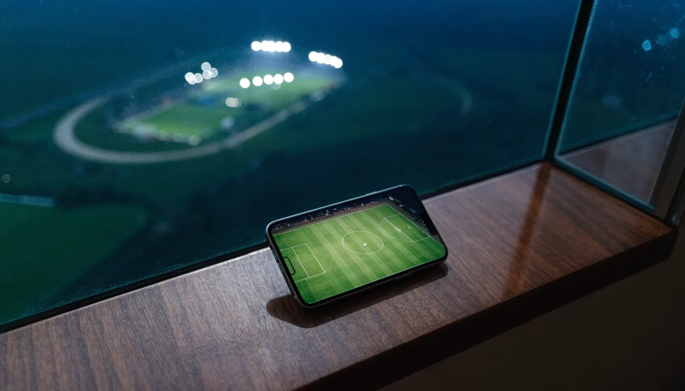 Ein Stadion unter Flutlicht von weitem gesehen, davor ein Feld und ein Feldweg im Dunkeln.